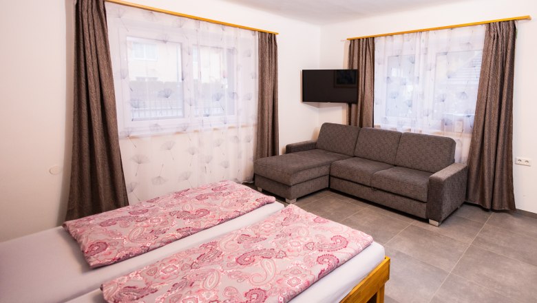Schlafzimmer, © Peter Prandsetter, Julian Eisenkirchner Ein Schlafzimmer mit Doppelbett, grauem Sofa, Fernseher und zwei Fenstern mit Vorhängen.