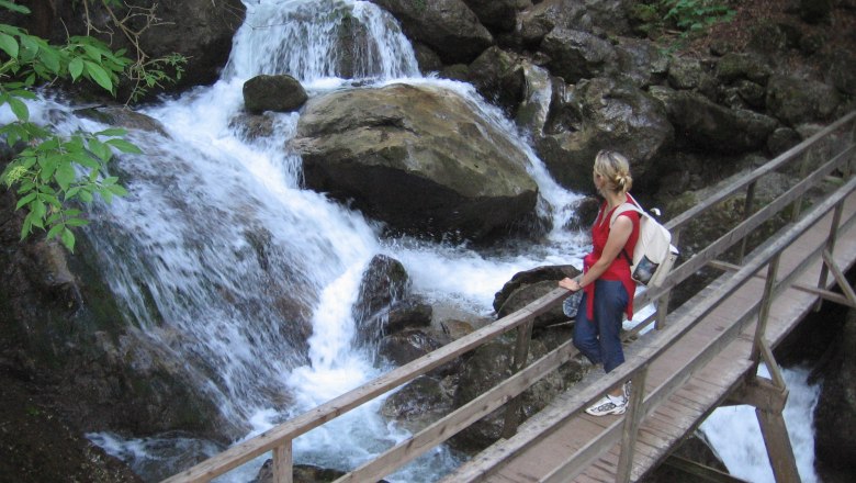 Myrafälle, © Roman Klementschitz, Wien Person auf Holzbrücke bei Wasserfall in bewaldeter Umgebung.