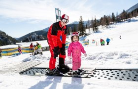 Am Fuße des Schneebergs Schifahren lernen, © NÖ Schneebergbahn, Franz Zwickl Ein Skilehrer hilft einem kleinen Kind beim Skifahren auf einem Förderband im Schnee.