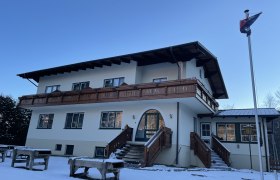 Frühstückspension Mariahilfberg, © Weiner Alpen Ein zweistöckiges Gebäude mit Holzbalkon und Treppen, umgeben von Schnee, unter klarem Himmel.