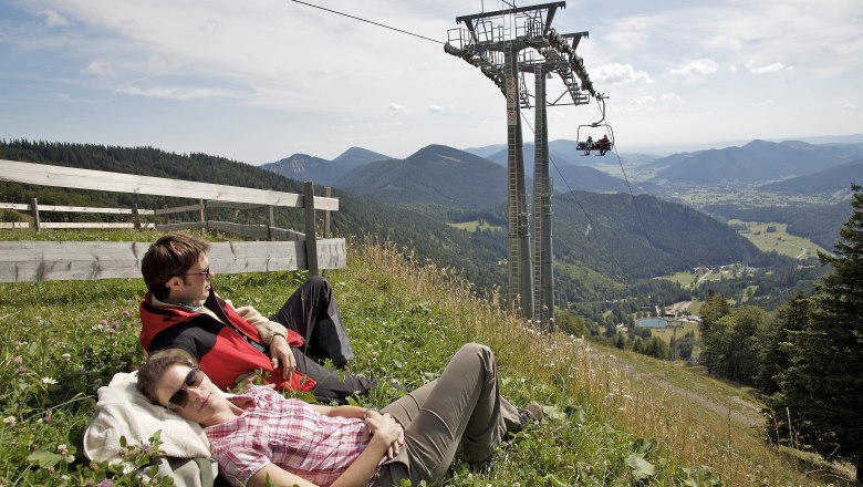 Schneeberg Sesselbahn, © NÖVOG/Franz Zwickl Zwei Personen entspannen auf einer Wiese neben einer Sesselbahn mit Bergblick.