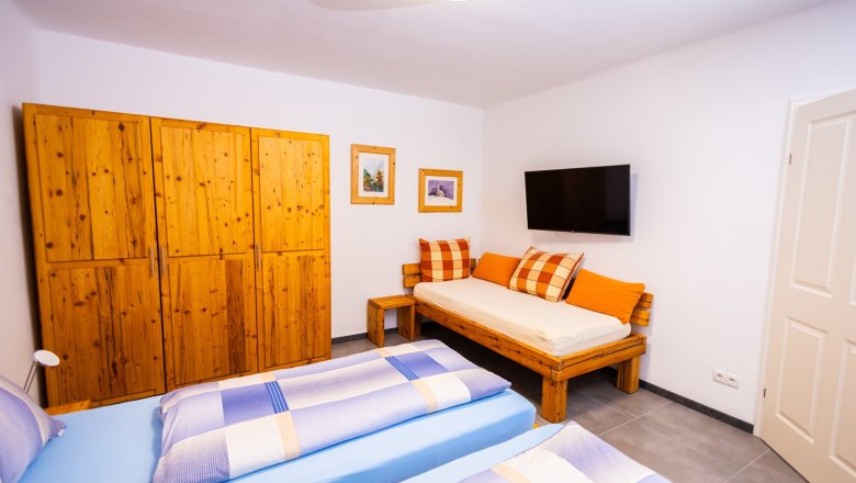 Schlafzimmer, © Peter Prandstetter, Julian Eisenkirchner Schlafzimmer mit Holzmöbeln, zwei Betten, Sofa, TV und Bildern an der Wand.