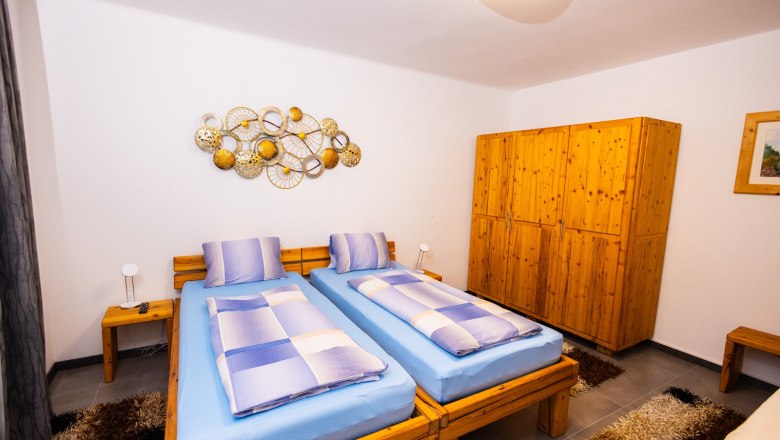 Schlafzimmer5(c)Peter Brandstetter, Julian Eisenkirchner, © Peter Prandstetter, Julian Eisenkirchner Ein gemütliches Schlafzimmer mit zwei Einzelbetten, blauer Bettwäsche, Holzmöbeln und dekorativer Wandkunst.