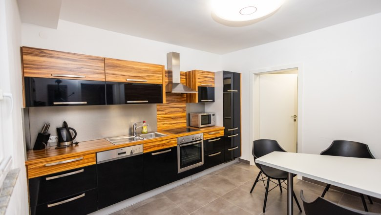 Küche, © Peter Prandstetter, Julian Eisenkirchner Moderne Küche mit Holz- und schwarzen Hochglanzfronten, Esstisch und Stühlen.