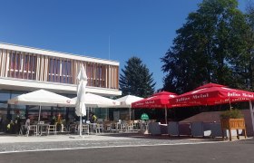 Der gemütliche Gastgarten des Hubertushofes, © Fotografin: Eva Heindl Ein moderner Gastgarten mit Sonnenschirmen und Sitzgelegenheiten vor einem Gebäude bei sonnigem Wetter.