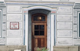 Eingang, © stein Eingangstür eines verzierten Gebäudes mit Holztür und dekorativen Malereien, umgeben von einem Holzzaun und Farnen.