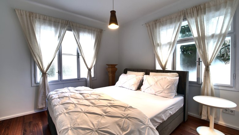 Schlafzimmer Top 2, © Daniela Sinadinoska Ein gemütliches Schlafzimmer mit einem großen Bett, weißen Vorhängen und einem kleinen runden Tisch.