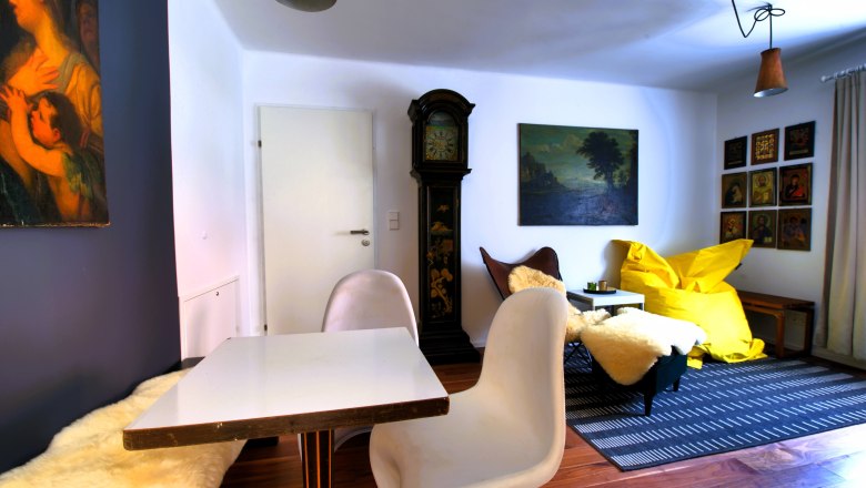 Wohnbereich Top 2, © Daniela Sinadinoska Wohnzimmer mit Tisch, Stühlen, Standuhr, Gemälden und gelbem Sitzsack.