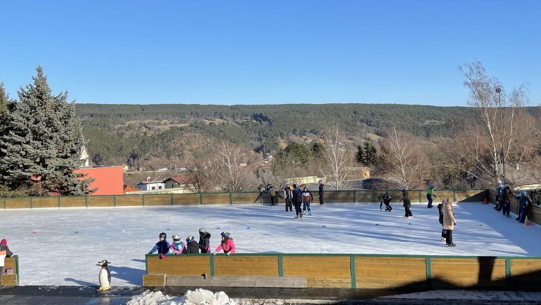 Eislaufplatz Piesting, © Wiener Alpen/Katharina Lechner Eislaufplatz mit Menschen, umgeben von Bäumen und Hügeln unter blauem Himmel.