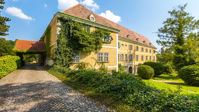 Schloss Strelzhof, © Wiener Alpen, Christian Kremsl Ein historisches Schloss mit gelber Fassade, bewachsen mit Efeu, umgeben von gepflegtem Garten und gepflastertem Weg.