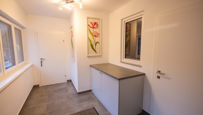 Vorraum, © Peter Prandstetter, Julian Eisenkirchner Moderner Vorraum mit weißen Wänden, grauem Boden, einem Schrank und Blumenbildern an der Wand.