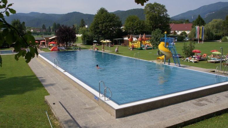 Ein Freibad mit Schwimmbecken, Rutsche und Liegewiese, umgeben von B&auml;umen und Bergen.