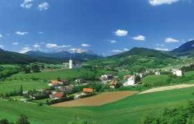Panoramablick auf H&ouml;flein mit Kirche, gr&uuml;nen Feldern und Bergen im Hintergrund.