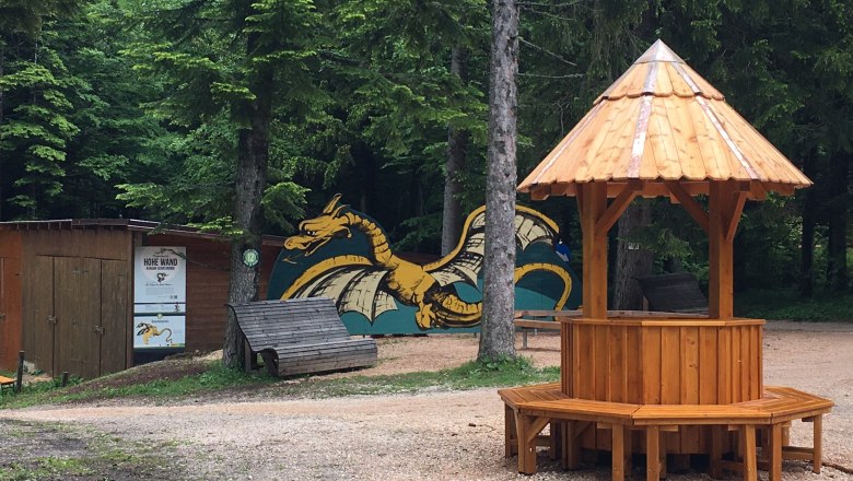 Ein h&ouml;lzerner Pavillon auf einem Waldplatz mit einem gemalten Drachen im Hintergrund.