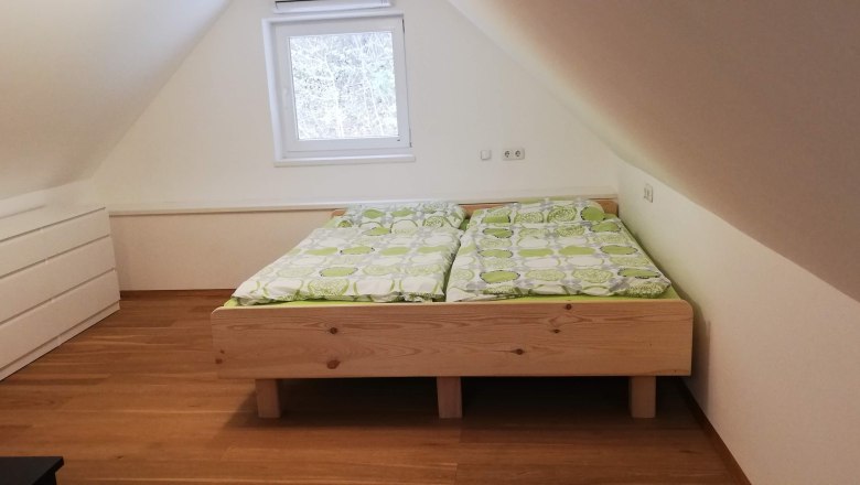 Ein Dachgeschosszimmer mit einem Doppelbett aus Holz und grüner Bettwäsche, daneben eine weiße Kommode.