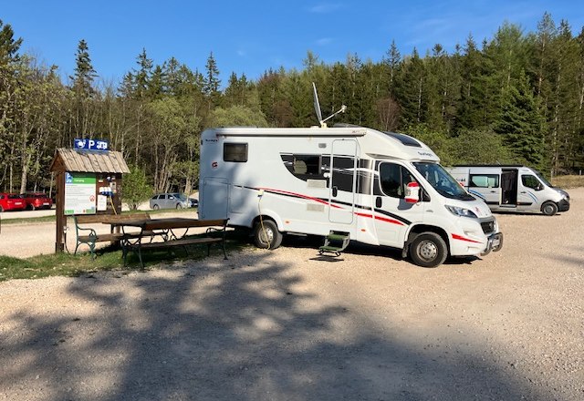 Wohnmobil auf einem Parkplatz im Naturpark Hohe Wand, umgeben von Bäumen und einem blauen Himmel.