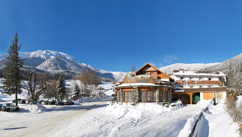 Winterlandschaft mit Hotel Forellenhof vor schneebedeckten Bergen und blauem Himmel.