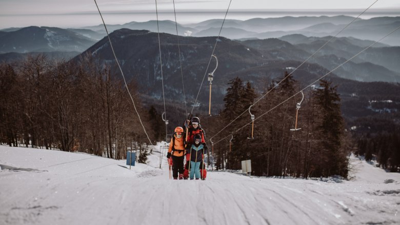 Drei Skifahrer auf einem Schlepplift im Schigebiet Unterberg, umgeben von schneebedeckten Bergen und Bäumen.