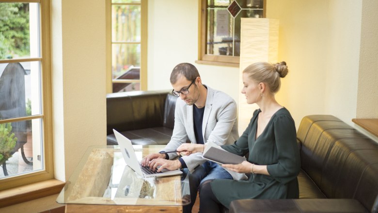 Zwei Personen arbeiten an einem Laptop in einem gemütlichen Raum mit großen Fenstern.