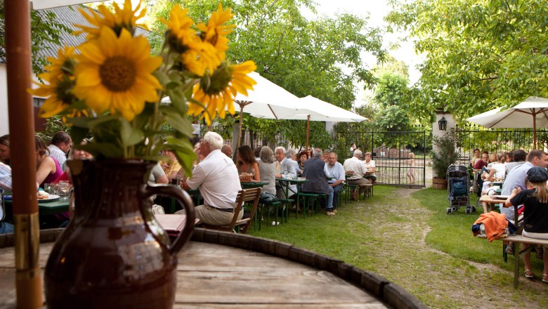 Ein Biergarten mit Menschen an Tischen, Sonnenschirmen und Sonnenblumen in einem Krug im Vordergrund.