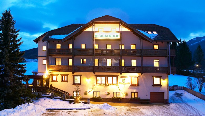 Beleuchtetes Geb&auml;ude der Pension Bruckerhof im Schnee bei D&auml;mmerung.