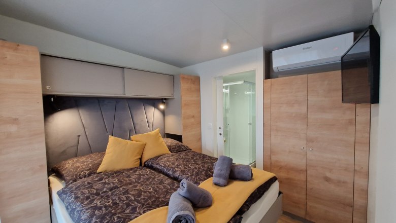 Innenansicht eines Schlafzimmers mit Doppelbett, gelben Kissen und Handtüchern, Schrank, Klimaanlage und Blick ins Badezimmer.