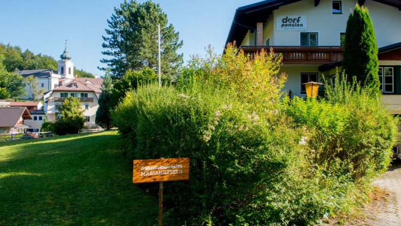 Ein Gebäude mit der Aufschrift 'Dorfpension' und einem Schild 'Frühstückspension Mariahilfberg' in einer grünen Umgebung.