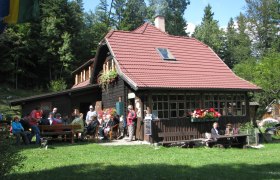 Eine Berghütte mit rotem Dach, umgeben von Wald, mit Menschen, die draußen sitzen.