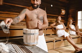 Mann gießt Wasser in Sauna, zwei Frauen sitzen entspannt.