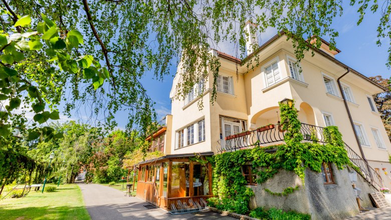 Ein gelbes Herrenhaus mit Efeu bewachsen, umgeben von Bäumen und einem Gartenweg.