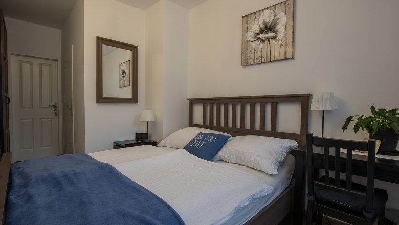 Gemütliches Doppelzimmer mit Holzbett, blauer Decke und Dekokissen. Ein Spiegel und ein Blumenbild an der Wand, daneben ein Schreibtisch mit Pflanze.