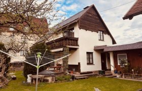 Ein zweist&ouml;ckiges Haus mit Balkon und Garten, umgeben von B&auml;umen und einem W&auml;schetrockner im Vordergrund.