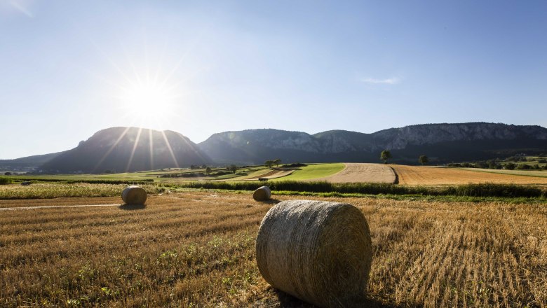 Landschaft mit Strohballen auf einem Feld, im Hintergrund Berge und die Sonne am Himmel.