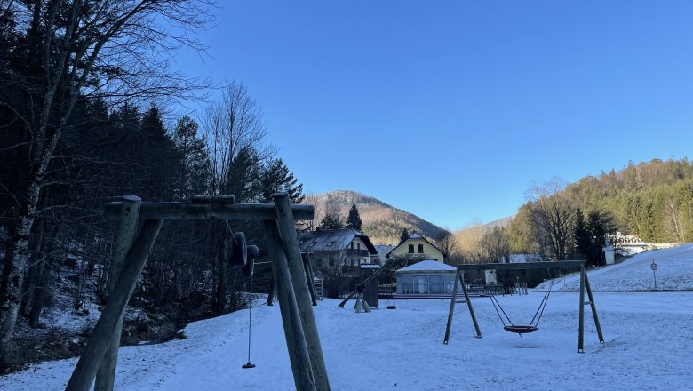 Winterlicher Spielplatz mit Schaukel und schneebedecktem Boden, umgeben von Bäumen und Bergen.