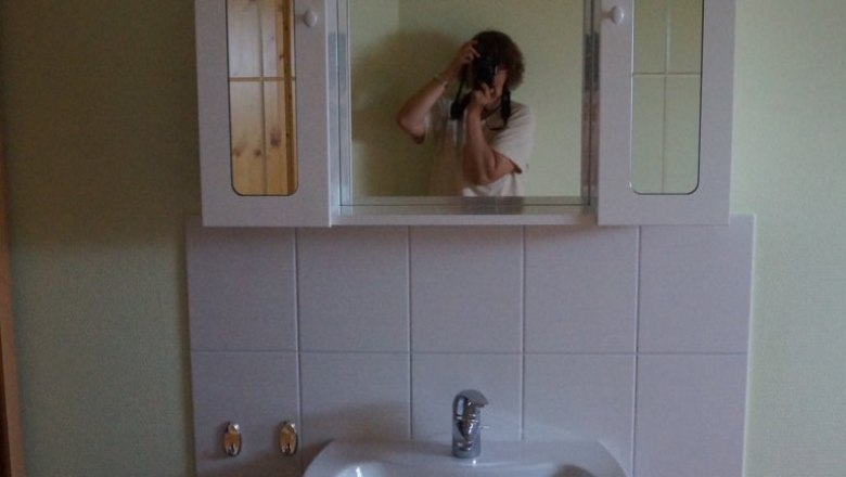 Ein Waschbecken mit Spiegel und Schrank in einem Zimmer. Eine Person fotografiert sich im Spiegel.