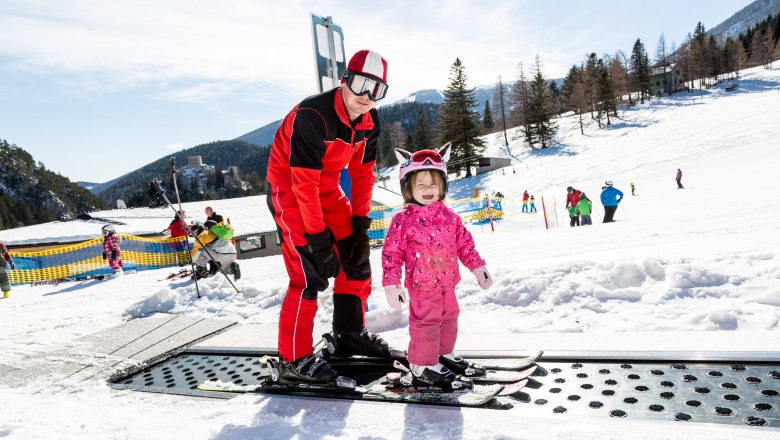 Ein Skilehrer hilft einem kleinen Kind beim Skifahren auf einem F&ouml;rderband im Schnee.