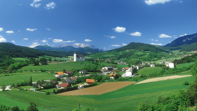 Panoramablick auf H&ouml;flein mit Kirche, gr&uuml;nen Feldern und Bergen im Hintergrund.