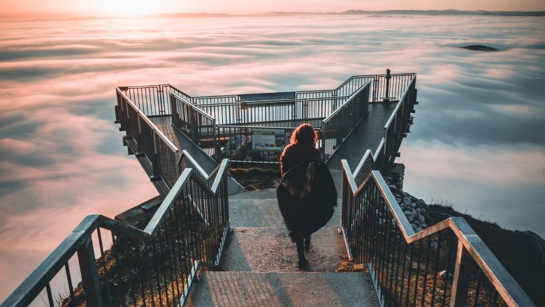 Eine Person geht auf einem Skywalk über einem Nebelmeer in den Sonnenuntergang.
