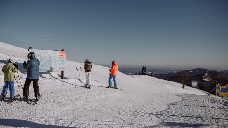 Skifahrer stehen auf einer Piste im Schigebiet Unterberg vor einem Wegweiser, umgeben von schneebedeckten Bergen und klarem Himmel.
