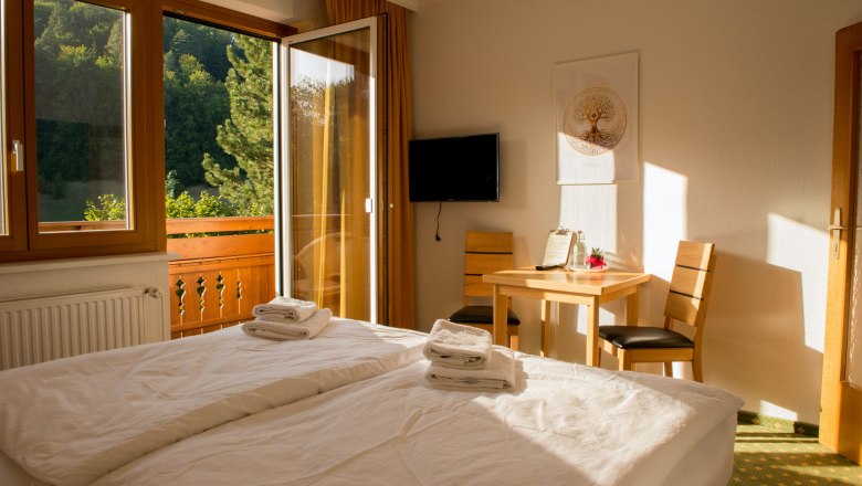 Ein helles Doppelzimmer mit Balkon, Bett, Tisch, Stühlen und einem Fernseher an der Wand. Durch die Balkontür sieht man grüne Bäume.
