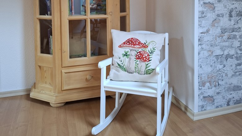 Ein Holzschrank mit B&uuml;chern und ein wei&szlig;er Schaukelstuhl mit einem Kissen, das Pilze zeigt, stehen in einem Raum mit Holzboden und gemusterter Tapete.