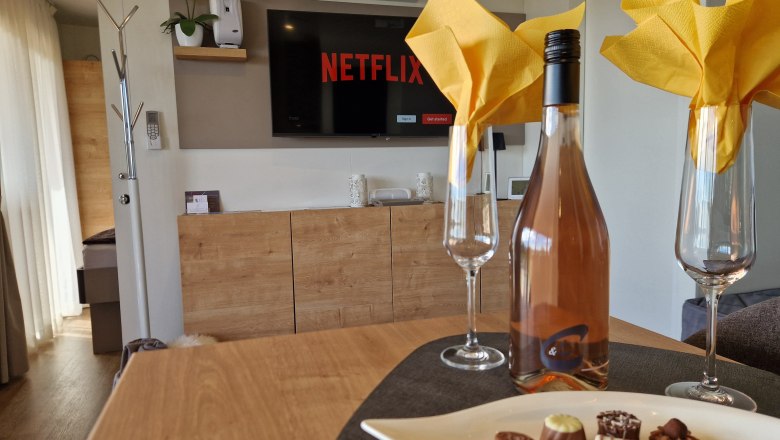 Innenansicht eines Chalets mit Weinflasche, Gläsern und Pralinen auf einem Tisch, Fernseher im Hintergrund.