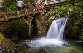 Holzbrücke über Wasserfall in bewaldeter Umgebung.