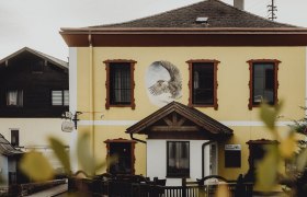 Gelbes Geb&auml;ude mit Wandgem&auml;lde eines Adlers und roten Fensterrahmen.