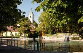 Thermalbad Bad Fischau-Brunn mit Kirche im Hintergrund, umgeben von Bäumen.