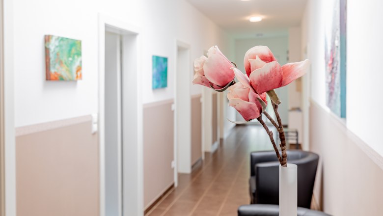 Ein heller Flur mit wei&szlig;en W&auml;nden, dekoriert mit Kunstwerken und einer Vase mit rosa Blumen im Vordergrund.