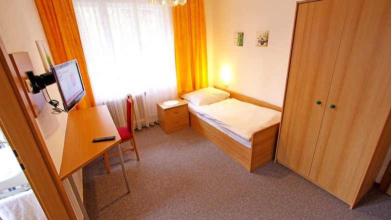 Ein einfach eingerichtetes Einzelzimmer mit Bett, Schreibtisch, Stuhl, Kleiderschrank und Fernseher. Gelbe Vorhänge und Teppichboden.