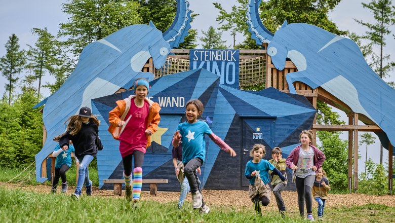 Kinder rennen vor einer blauen Holzkonstruktion mit Steinbock-Motiven und der Aufschrift 'Steinbock Kino'.