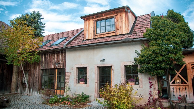 Rustikales Haus mit Ziegeldach und Holzverkleidung, umgeben von Pflanzen und Kopfsteinpflaster.