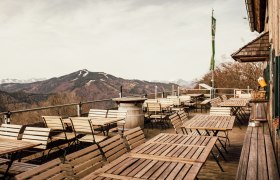 Leere Terrasse mit Holztischen und -st&uuml;hlen, Berglandschaft im Hintergrund.